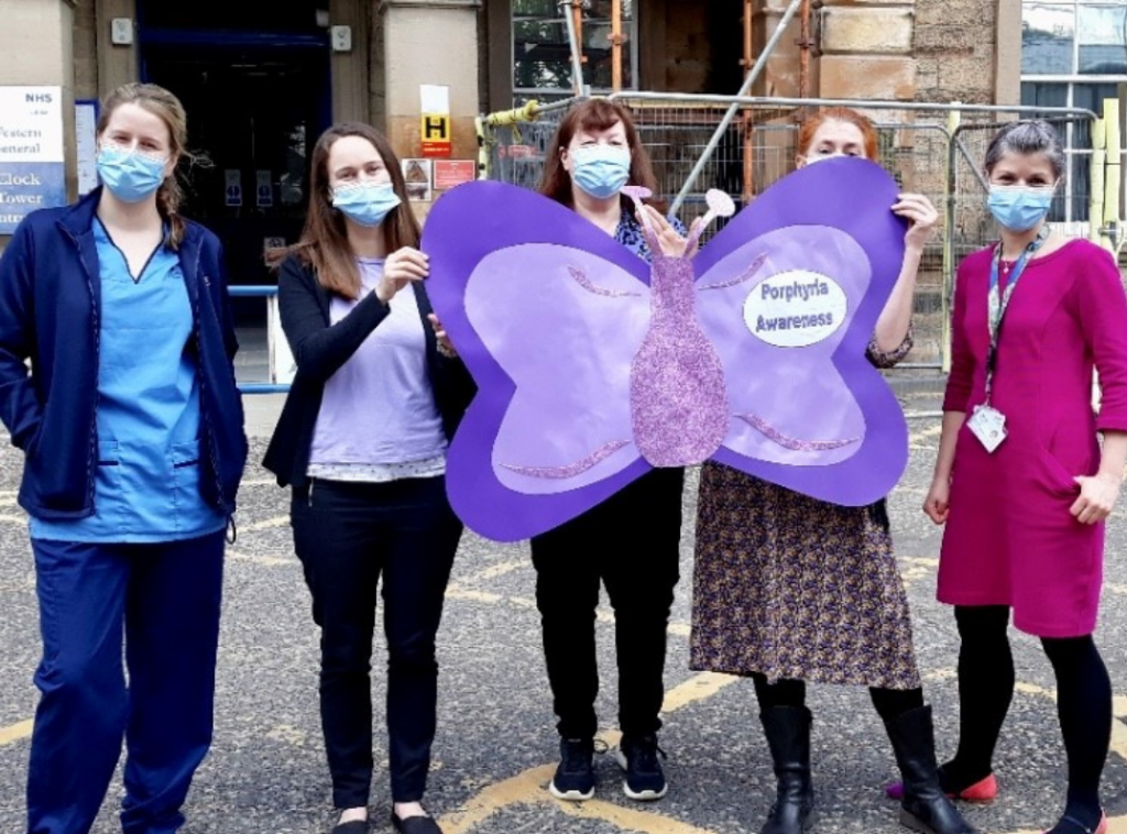 Photo of five white women are standing outside a hospital holding up a purple sign in the shape of a butterfly which reads "Porphyria Awareness". They are all wearing blue surgical face masks.