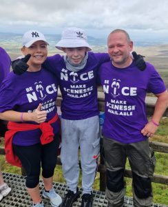 Oran, a teenage boy, stands in between his parents. Oran is wearing sun protective gear (hat, gloves, scarf) and they are all wearing purple t-shirts which read "NICE: Make Scenesse available". They are standing in front of a scenic mountain view.