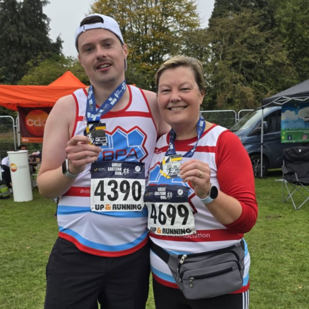 A man and a woman wearing BPA running vests are smiling and holding up medals after finishing a half marathon,